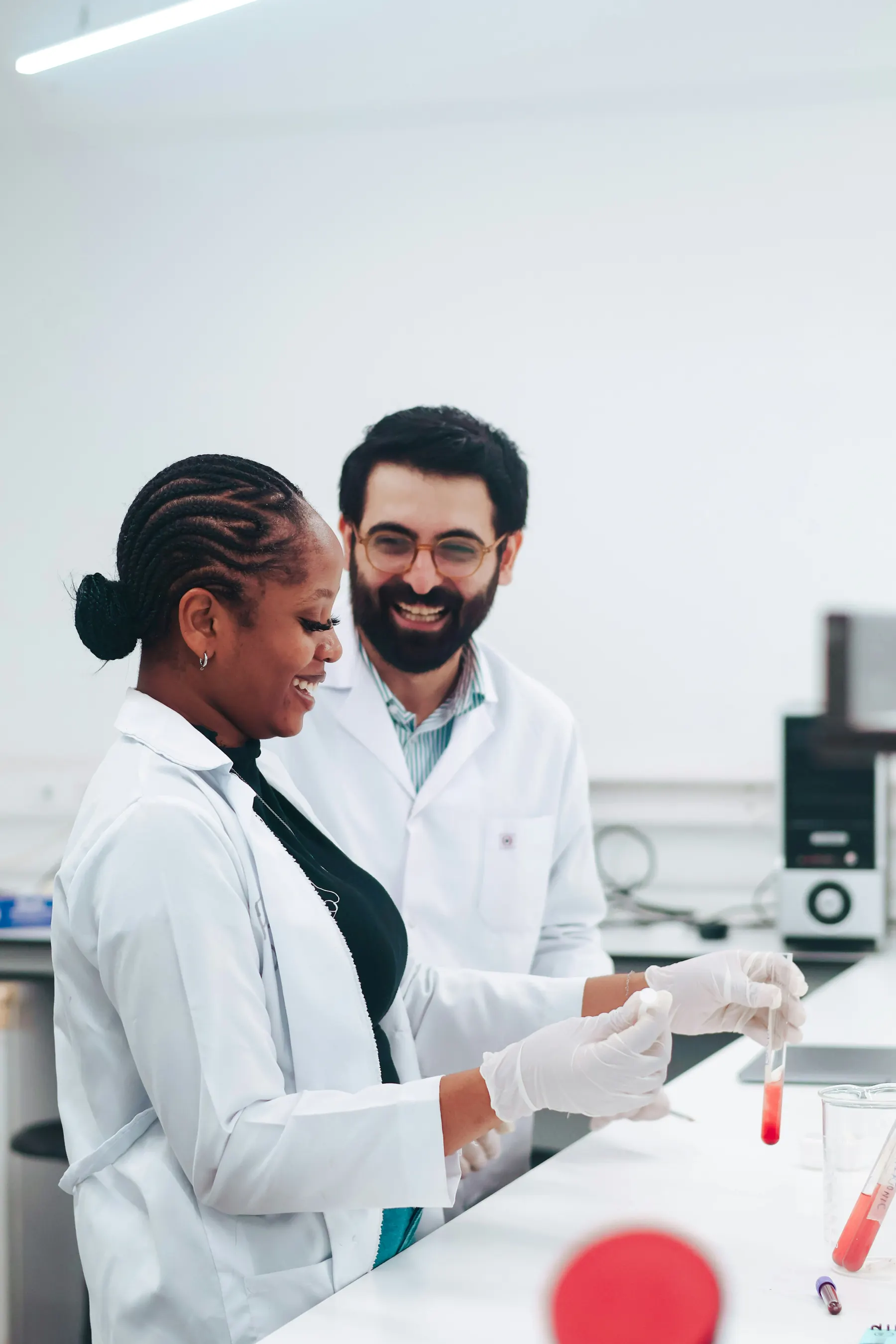 Two researchers in lab coats laughing together while examining a test tube sample